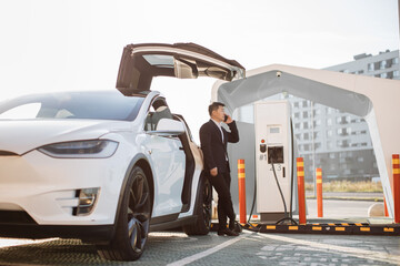 Positive asian man dressed in stylish business suit enjoying mobile conversation while waiting for his electric car charging battery. Modern EV station for innovative vehicle.