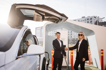 Happy caucasian businesswoman and asian man in black suit standing together near white electric car and shaking hands. Successful purchase of new luxury transport.