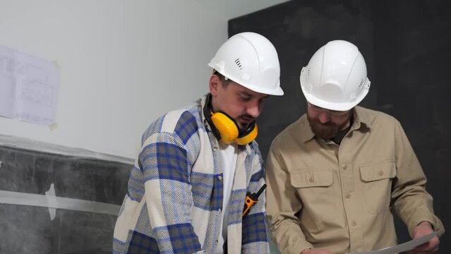 Two engineers in helmets at a construction site are leaning over a table with drawings and discussing them. Active discussion of construction documentation by two builders or architects. 4k footage