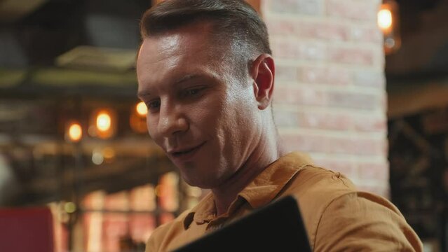 Tilt up shot of Caucasian man choosing dishes in menu and speaking with someone while sitting in restaurant