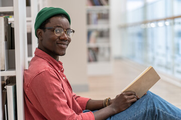 Black student man library visitor sitting on floor near bookcase with book in hands, looking at...