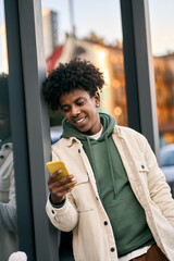 Cool smiling young African American teen guy holding mobile phone standing at glass city wall....
