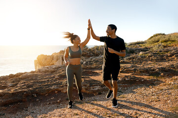 Smiling young african american male and female run in morning on rocks near ocean give high five