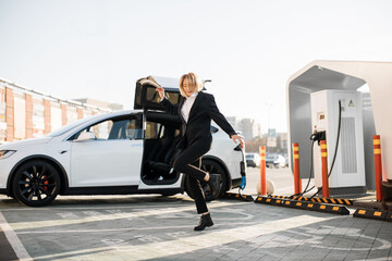 Charming young woman in business suit dancing joyfully outdoors while her luxury electro car charging on EV station. Enjoyment of free time with modern eco friendly transport.