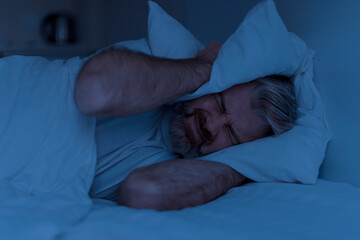 Closeup of angry mature man covering head with pillow