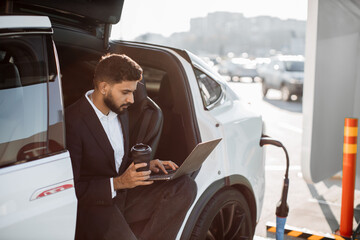 Focused man in business suit drinking coffee and working on laptop while charging battery of modern electro car. Concept of people, transport and technology.