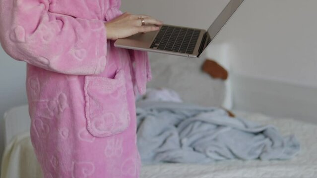 business woman in bathrobe working at home at laptop sitting on feet and baby playing in background lying on bed having fun.girl holding laptop in arms sliding with fingers on touchpad freelance