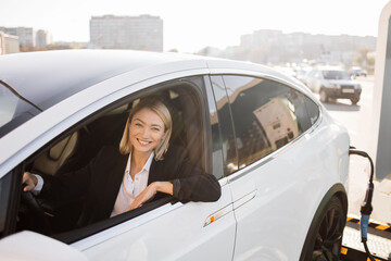 Caucasian woman sitting on driver seat in luxury car and smiling on camera through open window at EV station. Pleasant business lady with blond hair waiting for vehicle being refilled with energy.