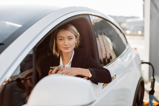Attractive caucasian woman with modern smartphone in hands sitting inside luxury car while recharging battery on auto station. Business lady in stylish suit working online during trip break.