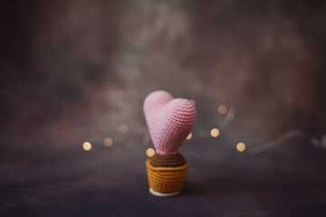 a pink knitted heart in a brown knitted pot is illuminated by the yellow light of a thin garland on a dark background