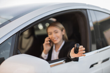 Happy female entrepreneur in suit sitting inside white luxury car and showing keys on camera from open window. Caucasian young blonde having mobile conversation in her new dream auto.