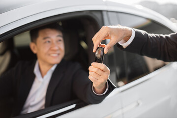 Happy male client of dealership in formal clothes receiving car keys from competent seller. Handsome asian businessman sitting inside new luxury auto with satisfied facial expression.