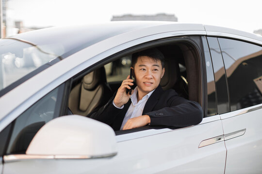Confident well-dressed businessman having pleasant conversation on mobile phone inside shiny new automobile. Focused asian male thinking over information heard from speaker while staring out window.