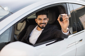 Fototapeta Handsome indian businessman sitting inside luxurious modern auto and demonstrating key out of open car window. Cheerful smiling young guy in smart suit enjoying new auto at driver seat. obraz