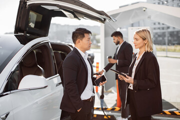 Focused multiethnic coworkers in formal clothing holding modern gadgets in hands. Arabic colleague with takeaway coffee supplying battery-drive vehicle with energy at public charging point.