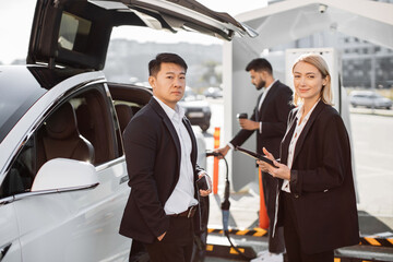 Relaxed smiling multiracial business colleagues standing at e-car charging point with tablet and mobile in hands. Indian man holding takeaway cup and getting auto filled up with electricity behind.