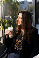 Smiling, charming woman holding a drink in her hand, smiling and looking away on the street. A dreamy girl during a lunch break.