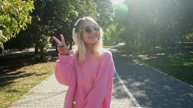 A teenager girl in pink glasses dances and shows a victory sign with her hand, a sunny day and a good mood, listens to music in headphones