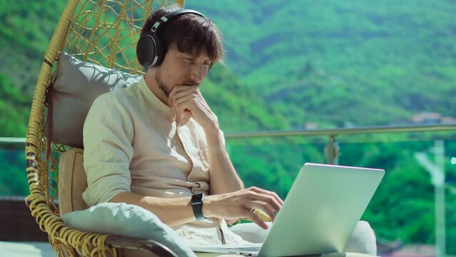 Young man on balcony, surrounded by mountain scenery. Wearing headphones, focused on laptop screen. Searching for mistake solution. Perfect for tech, problem-solving, or remote work projects. 