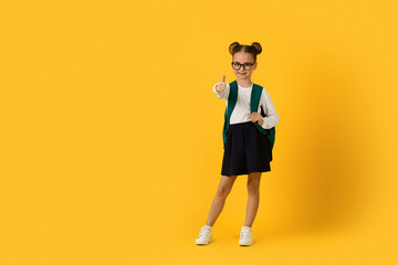 Smiling Cute Schoolgirl Gesturing Thumb Up While Standing Over Yellow Studio Background