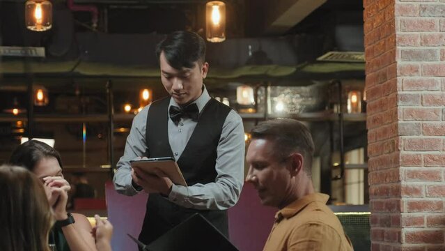 Tilt up shot of Asian waiter in elegant uniform using digital tablet while taking order from family with kid in restaurant