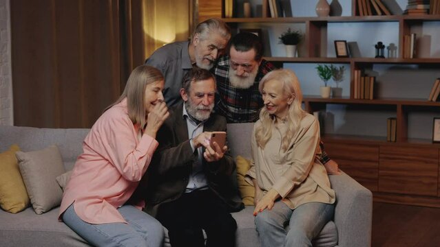 Group of Happy Senior Friends Using Smartphone Sitting on Sofa in Nursing Home. Mature Man Showing Something to His Friends, Scrolling Photos. Old People Spending Time Together
