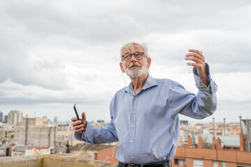 Mature businessman with gray hair and beard in blue shirt, jeans and glasses listen to music with earphones and dancing on rooftop terrace with city skylight on background. Copy space