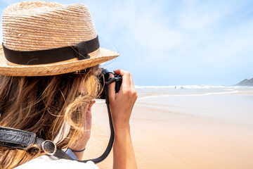 A young woman in a straw hat with windswept blonde hair takes photos of distant surfers on the wide...