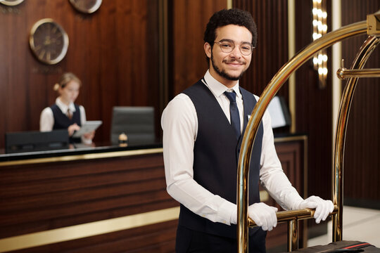 Young elegant bellboy in uniform pushing cart with clients baggage while moving along hotel lounge and looking at camera
