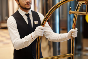 Close-up of elegant bellboy in uniform and white gloves pushing cart with baggage of tourists while moving along lobby in front of camera © pressmaster