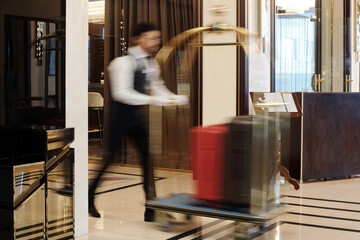 Blurry motion of bellboy walking fast along lounge or hall of luxurious five star hotel and pushing...
