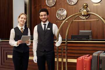Happy young elegant receptionist and bellboy standing in lounge of luxurious hotel and looking at camera while waiting for new guests - obrazy, fototapety, plakaty
