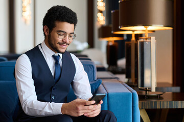 Happy young broker in elegant suit texting in mobile phone or communicating in video chat while...