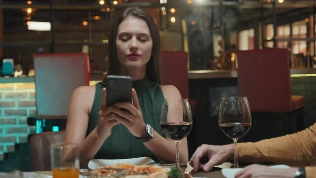 Medium shot of female blogger photographing food with smartphone while having dinner in restaurant