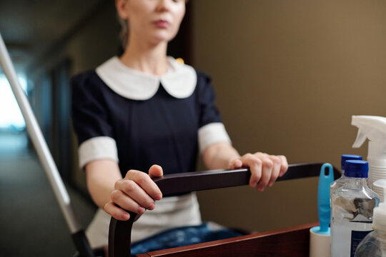 Close-up of young chambermaid pushing cart with detergents in bottles while moving along corridor in hotel and during room service