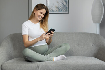 Smiling pregnant woman holding mobile phone, reading text message, shopping online sitting on sofa at home. Pregnancy concept 