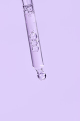 Facial serum in glass pipette on light purple background. Liquid gel or essential oil with bubbles macro. Trendy anti aging cosmetic product with hyaluronic acid closeup. Copy space, front view.