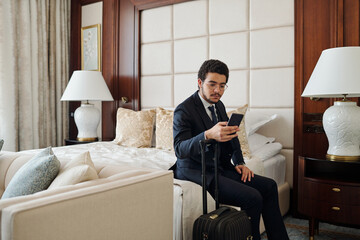 Young serious businessman in formalwear reading message in smartphone while sitting on double bed...