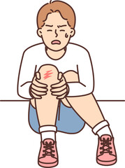 Crying boy sits on ground and holds on to injury on knee after fall during walk
