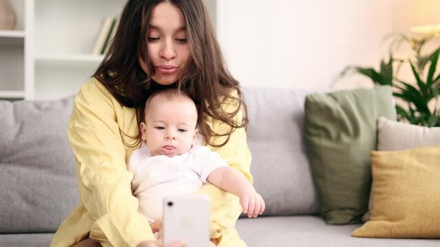Portrait of cute young woman mother hug her little baby and grimacing taking selfie photograph saving great memories on smartphone mobile phone at light home Happy motherhood concept