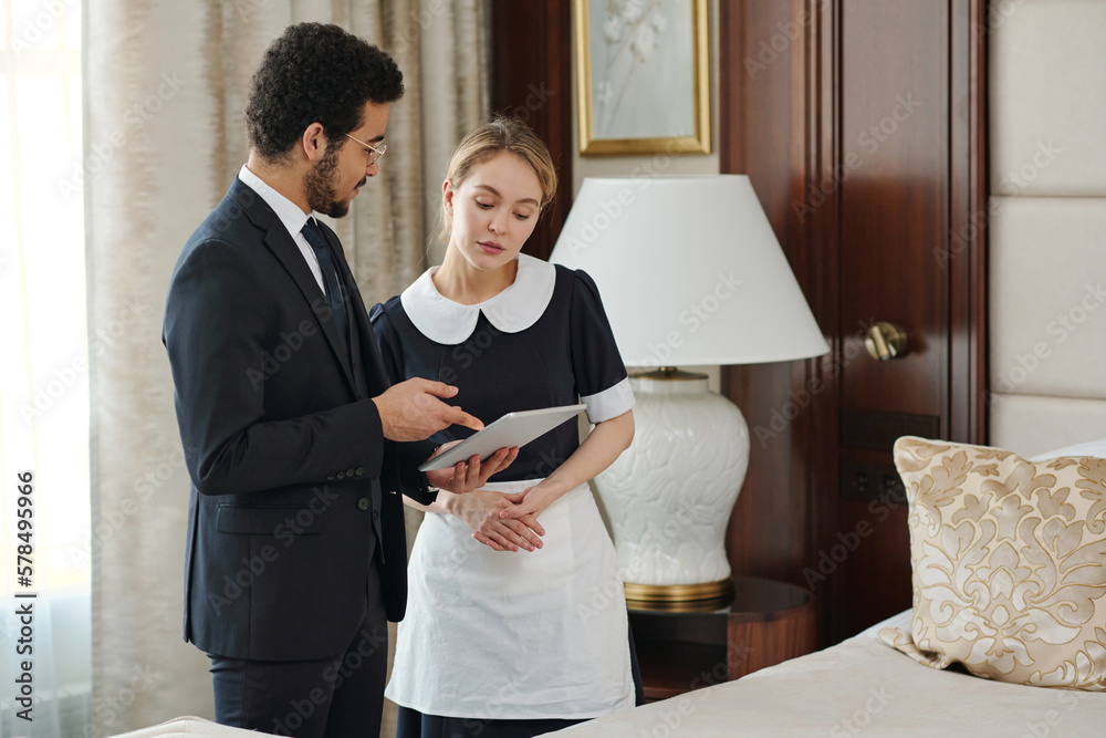 Poster confident owner of luxurious hotel giving instructions to chambermaid while pointing at tablet scree