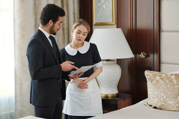 Confident owner of luxurious hotel giving instructions to chambermaid while pointing at tablet...