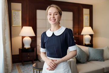 Young successful chambermaid in uniform standing in front of camera against couch and two lamps in...