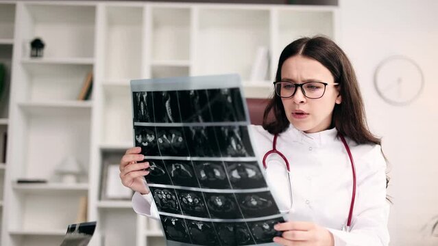 Portrait of concentrated young woman doctor therapist with glasses thinking about diagnosis while looking at results patient MRI or CT scan procedure at light hospital office