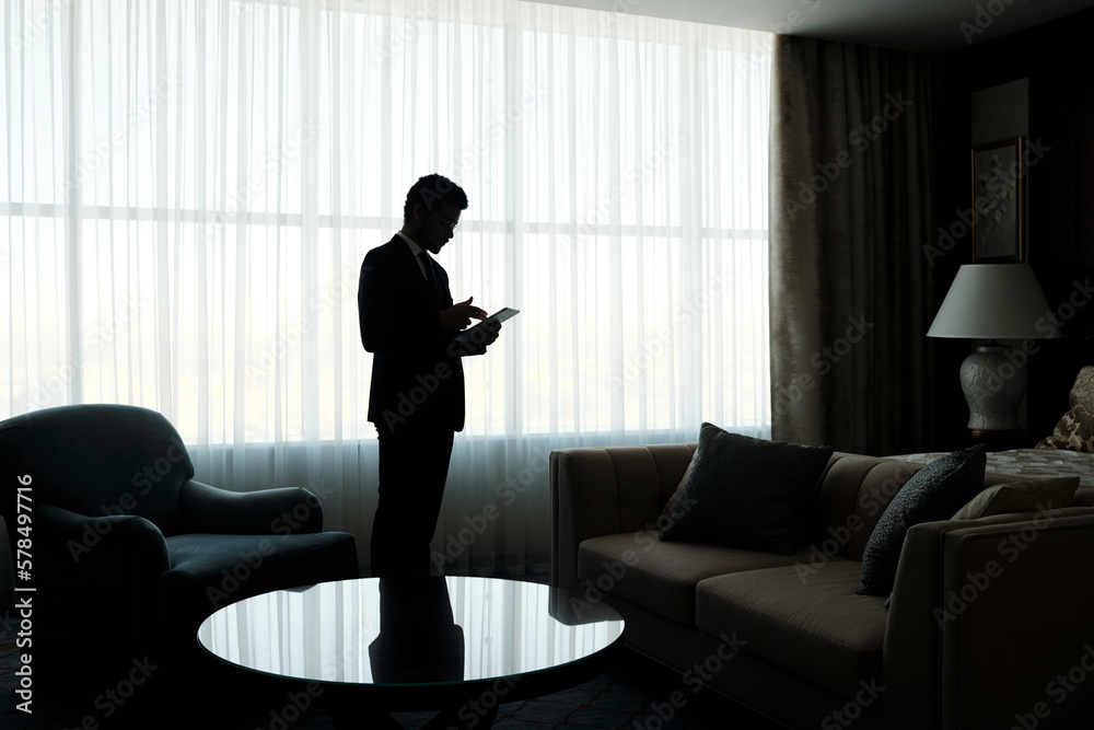 Poster outline of young businessman using tablet while standing against large window covered with white chi
