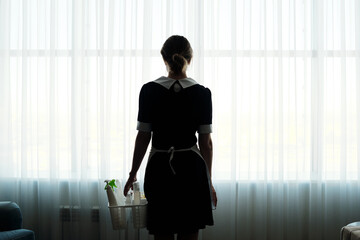 Fototapeta na wymiar Rear view of chambermaid with detergents in basket walking towards large window covered with white chiffon curtains in hotel room