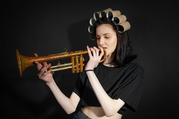 Portrait of a poor, skinny girl in black, rumpled clothes and a strange hat that made of toilet paper tubes holding a rusty trumpet, isolated on a black background.