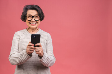 Happy mature senior woman holding smartphone using mobile online apps, smiling old middle aged grandmother texting sms message chatting on phone isolated over pink background. - 578498381