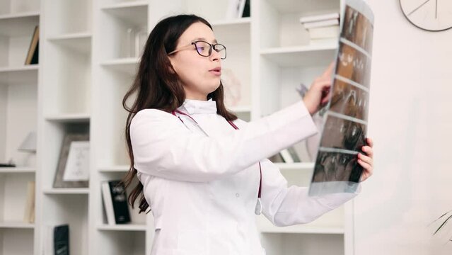 Portrait of positive young woman doctor therapist with glasses looking at great good results patient MRI or CT scan procedure and looking at camera with thumbs up at light hospital office
