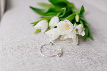 Bracelet in white gold lies inext to a bouquet of white tulips on white background. Gift for Women's Day, Valentine's Day. Beautiful spring background with green leaves. Mock up